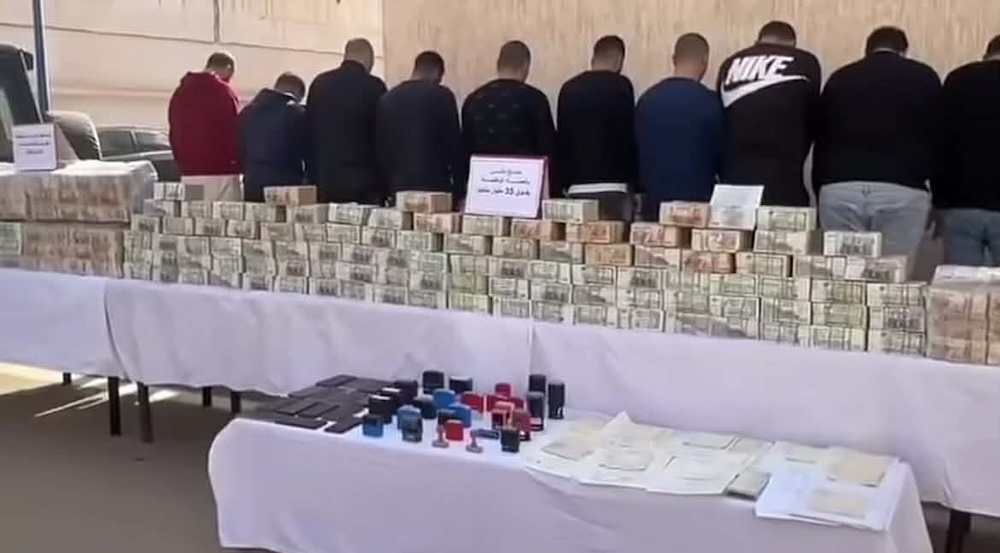 Des suspects alignés de dos devant des tables présentant des liasses de billets de banque et du matériel saisi lors d'une opération de police contre un réseau de fraude.